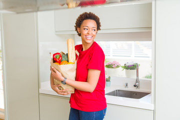 Young beautiful african american woman holding paper bag full of fresh healthy groceries at the kitchen