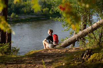 Fototapeta na wymiar Caucasian short hairstyle woman with red backpack sitting on a log above river and looking faraway. Relaxation.