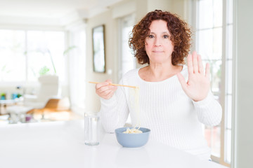 Senior woman eating asian noodles using chopsticks with open hand doing stop sign with serious and confident expression, defense gesture