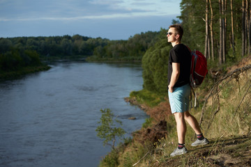 Fototapeta na wymiar Caucasian young man with red travel backpack standing on the brink above forest river in summer and looking faraway