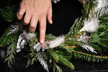 Female hand is holding silver ginkgo leaf to glue it into winter wreath. DIY Christmas wreath decorations with fir tree branches and silver painted leaves and white feathers. Handmade wreath crafting.