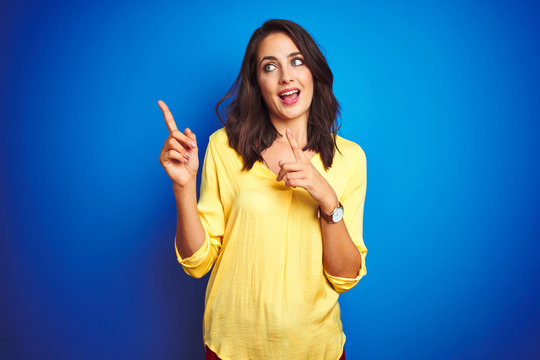 Young beautiful woman wearing yellow t-shirt standing over blue isolated background smiling and looking at the camera pointing with two hands and fingers to the side.