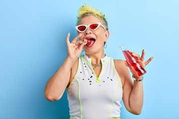 glamour charming mature woman sucking, licking lollipop, isolated over blue background, studio shot.