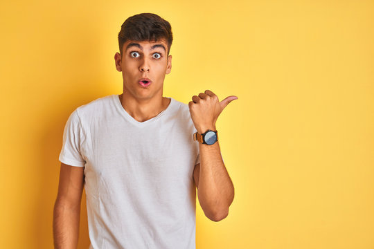 Young indian man wearing white t-shirt standing over isolated yellow background Surprised pointing with hand finger to the side, open mouth amazed expression.