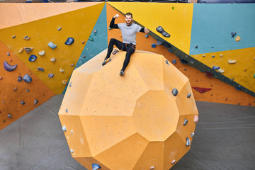 Young cheerful physically challenged boulderer sitting on top of large round-shape artificial rock wall, showing his biceps, being absolutely happy and proud of himself, glad to reach high results.