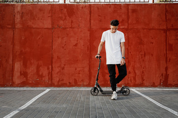 Young guy dressed in jeans and t-shirt stands with a scooter against a painted concrete wall on the summer day in the city. © Leika production