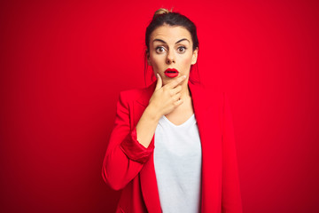 Young beautiful business woman standing over red isolated background Looking fascinated with disbelief, surprise and amazed expression with hands on chin