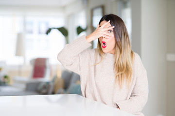 Young beautiful woman wearing winter sweater at home peeking in shock covering face and eyes with hand, looking through fingers with embarrassed expression.