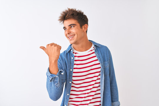Young handsome man wearing striped t-shirt and denim shirt over isolated white background smiling with happy face looking and pointing to the side with thumb up.