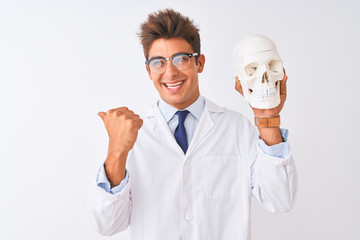 Young handsome doctor man wearing glasses holding skull over isolated white background pointing and showing with thumb up to the side with happy face smiling