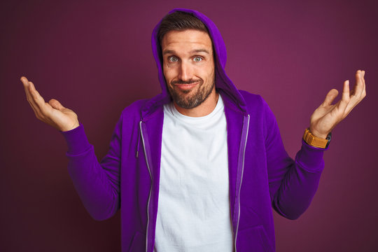 Young fitness man wearing casual sports sweatshirt and hood over purple isolated background clueless and confused expression with arms and hands raised. Doubt concept.