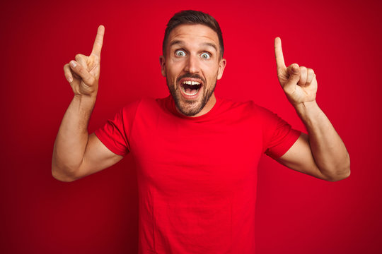 Young handsome man wearing casual t-shirt over red isolated background smiling amazed and surprised and pointing up with fingers and raised arms.