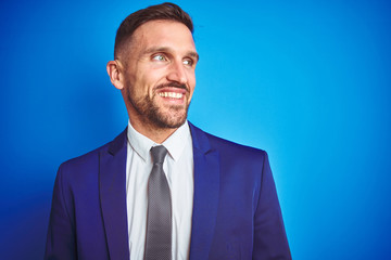 Close up picture of young handsome business man over blue isolated background looking away to side with smile on face, natural expression. Laughing confident.