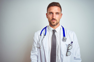 Young handsome doctor man wearing white professional coat over isolated background with serious expression on face. Simple and natural looking at the camera.