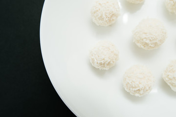 Chocolate covered coconut candies on white plate isolated on black background. Truffle candy set. Selective focus