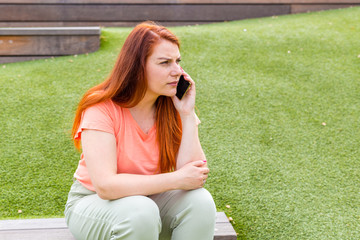 Dissatisfied young woman calling by smart phone outdoors, girl sitting on a bench in the park.