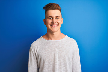 Young handsome man wearing white casual t-shirt standing over isolated blue background with a happy face standing and smiling with a confident smile showing teeth