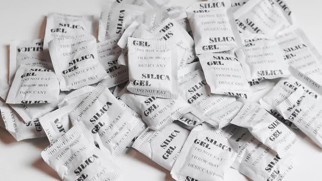 Pile of silica gel packets on a white table, with camera looking into the centre. Silica gel packets are used to absorb moisture and keep things dry.