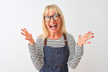 Middle age woman wearing apron and glasses standing over isolated white background celebrating crazy and amazed for success with arms raised and open eyes screaming excited. Winner concept