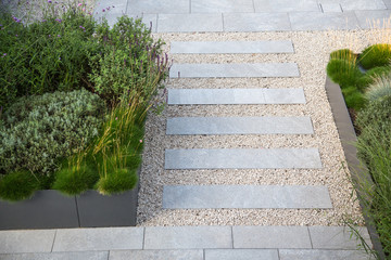 Moderne Garten- und Terrassengestaltung im Materialmix: Terrasse und Gehweg aus Steinplatten umgeben von Schotter und Metall Pflanzgefäßen mit Grünpflanzen 