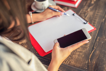 A woman's hands are writing on notebook and using smartphone on wooden table at cafe.