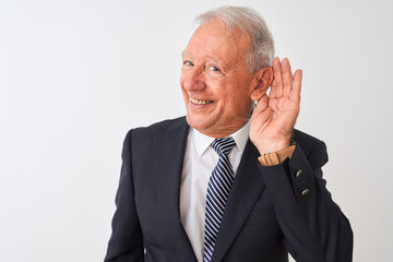 Senior grey-haired businessman wearing suit standing over isolated white background smiling with hand over ear listening an hearing to rumor or gossip. Deafness concept.