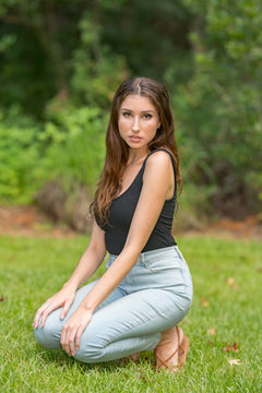 Attractive woman squatting in a park scene wearing tank top and denim jeans