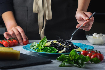 The girl winds black spaghetti on a fork. Unusual cuttlefish ink pasta. A wholesome light dinner with basil and parmesan.