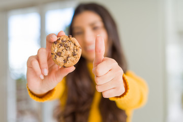 Young woman eating chocolate chips cookies at home happy with big smile doing ok sign, thumb up with fingers, excellent sign