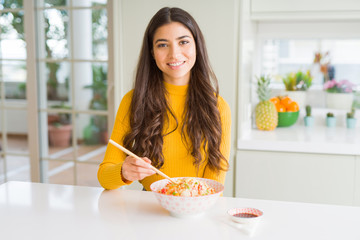 Young woman eating a bowl of Asian rice using chopsticks with a happy face standing and smiling...