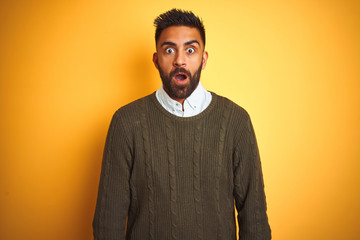 Young indian man wearing green sweater and shirt standing over isolated yellow background afraid and shocked with surprise expression, fear and excited face.