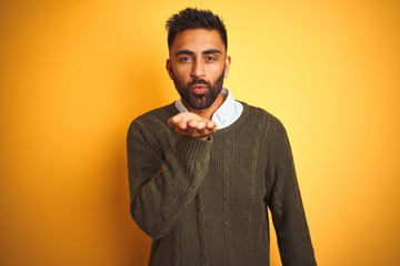 Obraz premium Young indian man wearing green sweater and shirt standing over isolated yellow background looking at the camera blowing a kiss with hand on air being lovely and sexy. Love expression.