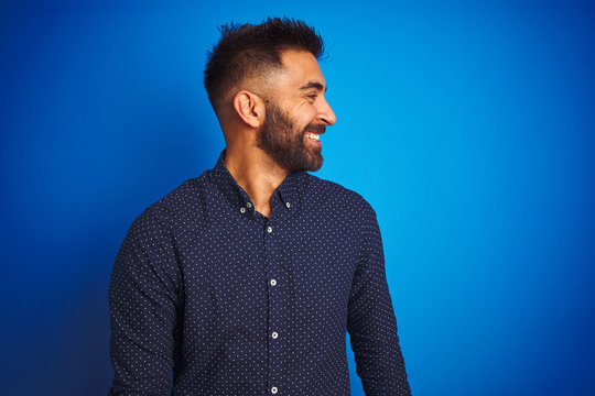 Young indian elegant man wearing shirt standing over isolated blue background looking away to side with smile on face, natural expression. Laughing confident.
