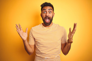 Young indian man wearing t-shirt standing over isolated yellow background celebrating crazy and...