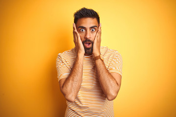 Young indian man wearing t-shirt standing over isolated yellow background afraid and shocked, surprise and amazed expression with hands on face - obrazy, fototapety, plakaty