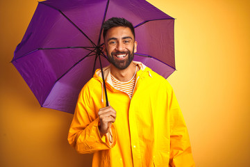 Young indian man wearing raincoat and purple umbrella over isolated yellow background with a happy...