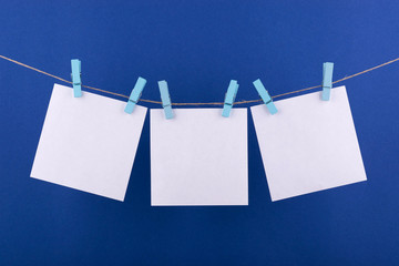 notepad paper sheets for notes and reminders of white color, fastened with decorative orange clothespins hanging on a rope on a blue background