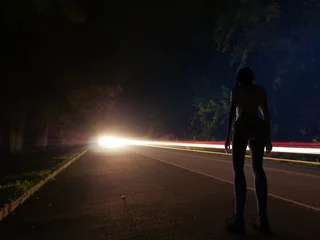 Tuinposter A young woman or girl all alone standing beside a road at night after dark with a long exposure background of car lights streaking by in a forest deep in the secluded hills and woods of Tennessee. © Michael