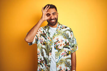 Fototapeta premium Young indian man on vacation wearing summer floral shirt over isolated yellow background doing ok gesture with hand smiling, eye looking through fingers with happy face.