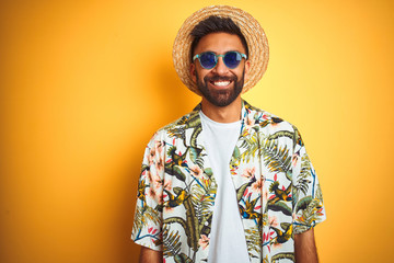 Indian man on vacation wearing floral shirt hat sunglasses over isolated yellow background with a...