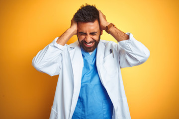 Young indian doctor man standing over isolated yellow background suffering from headache desperate and stressed because pain and migraine. Hands on head.