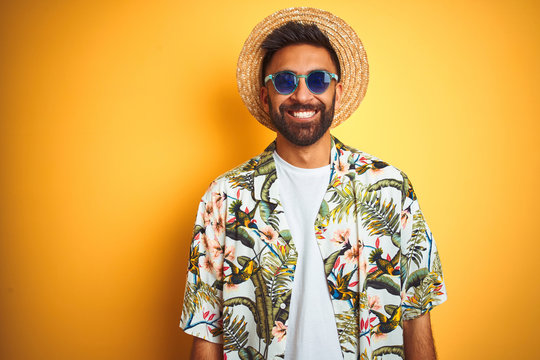 Indian man on vacation wearing floral shirt hat sunglasses over isolated yellow background with a happy and cool smile on face. Lucky person.
