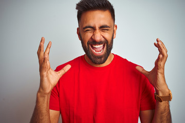 Young indian man wearing red t-shirt over isolated white background celebrating mad and crazy for success with arms raised and closed eyes screaming excited. Winner concept