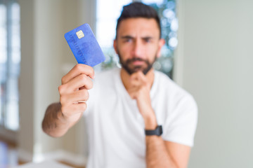 Handsome hispanic man holding credit card serious face thinking about question, very confused idea