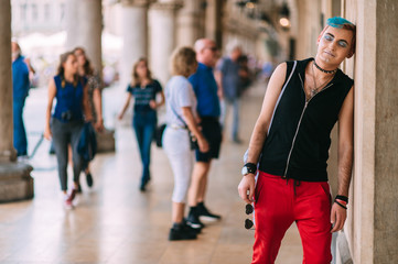 Transgender man in stylish clothing with makeup and colored hairstyle lifestyle portrait in the city with people on background.