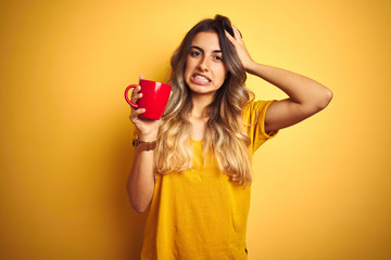 Young beautiful woman holding  red cup of coffee over yellow isolated background stressed with hand on head, shocked with shame and surprise face, angry and frustrated. Fear and upset for mistake.