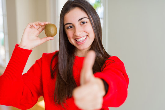 Beautiful young woman eating fresh green kiwi happy with big smile doing ok sign, thumb up with fingers, excellent sign