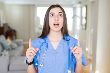 Beautiful young nurse woman wearing uniform and stethoscope at the clinic amazed and surprised looking up and pointing with fingers and raised arms.