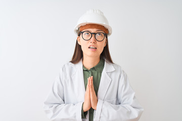 Young chinese engineer woman wearing coat helmet glasses over isolated white background begging and...
