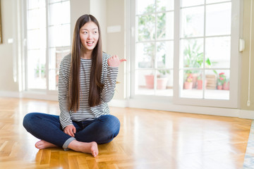 Beautiful Asian woman sitting barefoot on the floor at home smiling with happy face looking and pointing to the side with thumb up.